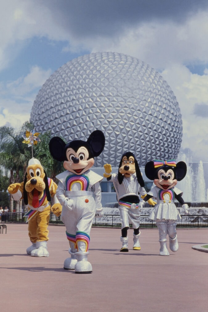mickey, minnie, goofy and pluto costumed characters walk in front of the large silver epcot sphere