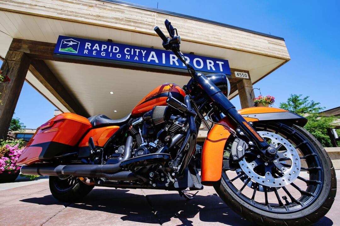 Close-up from below of an orange motorcycle with a sign behind it reading "Rapid City Regional Airport"