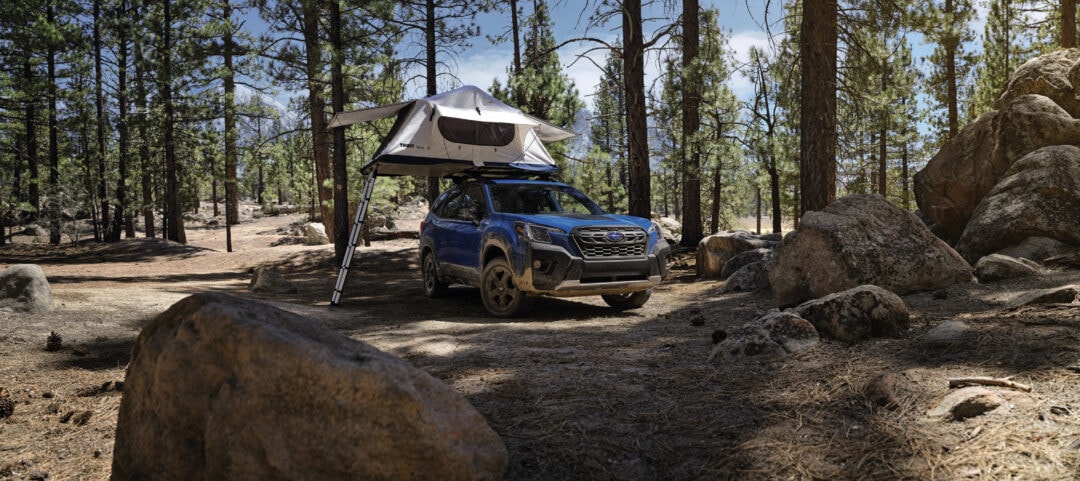a blue subaru with a pop up tent parked in the woods near some large rocks