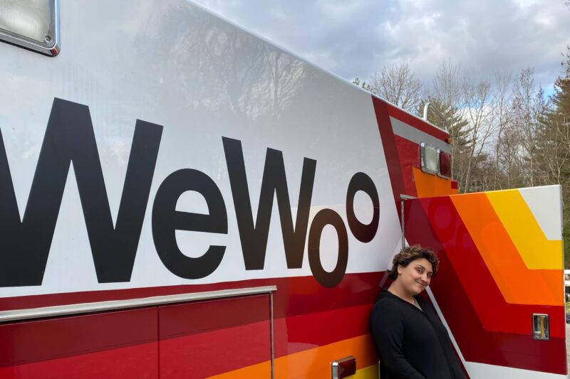 a person stands next to an ambulance painted with red, orange, and yellow stripes and the word "WeWoo"