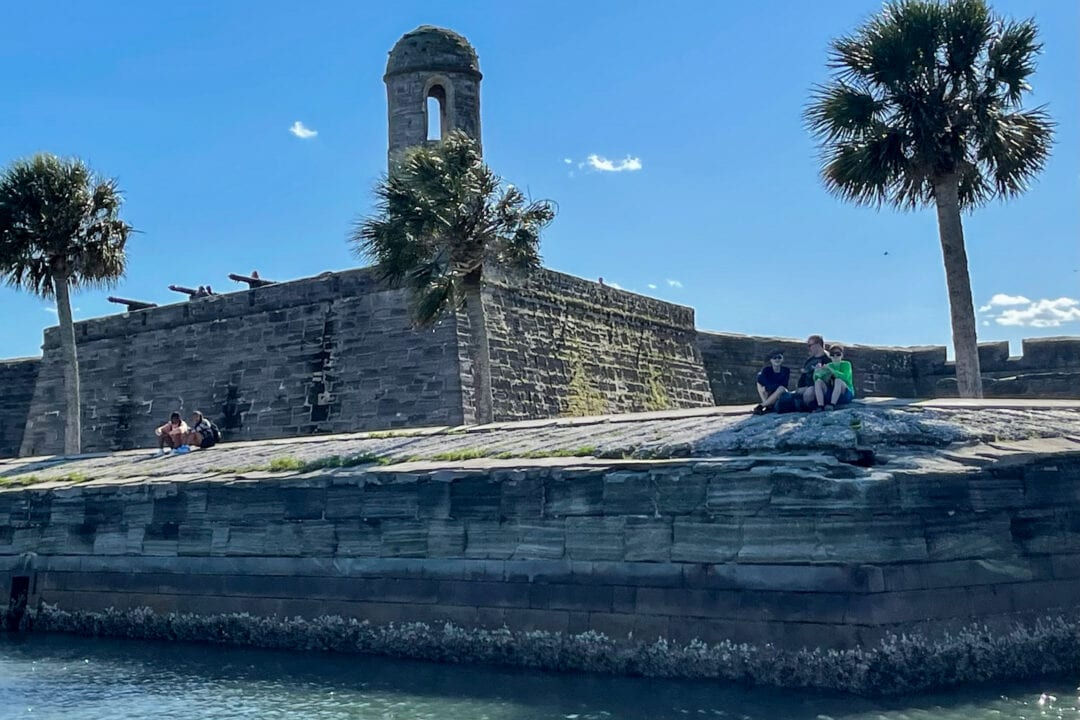 a few people sit on a stone fort by the water with palm trees