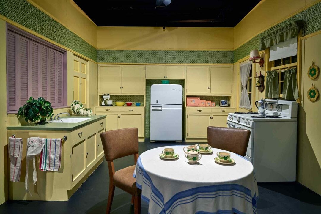 a 1950s kitchen set with yellow and green walls, a white fridge and stove, and a small circular kitchen table