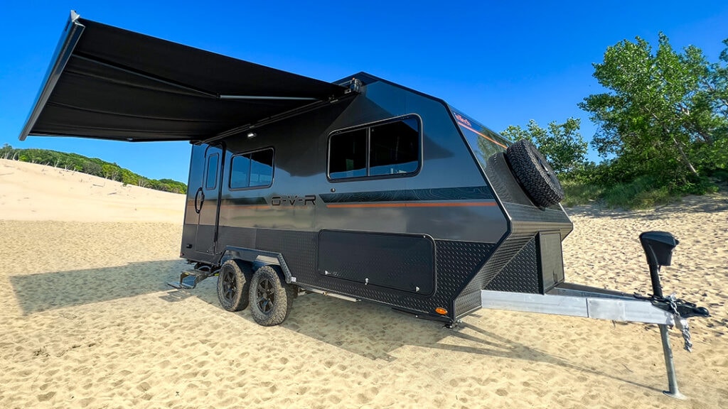 A rugged black travel trailer with its awning out, parked on a sandy beach
