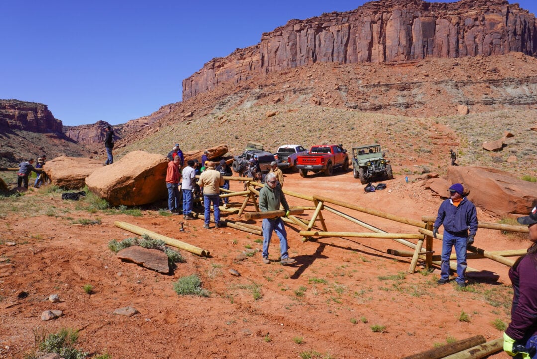 A group of people in a red rock desert landscape carrying logs and building a fence
