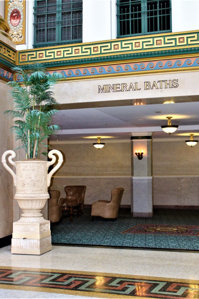 an ornate hotel lobby with a plant in a large marble urn, decorative yellow, green and blue trim, and gold leaf lettering that says "mineral baths"