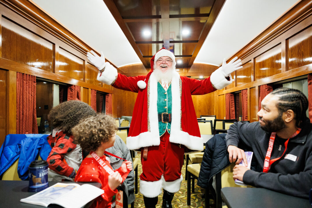 Santa stands with outstretched arms as he greets passengers on a train.