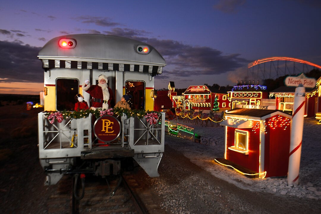 Santa waves from the caboose of a train as it passes an illuminated town marked "North Pole."