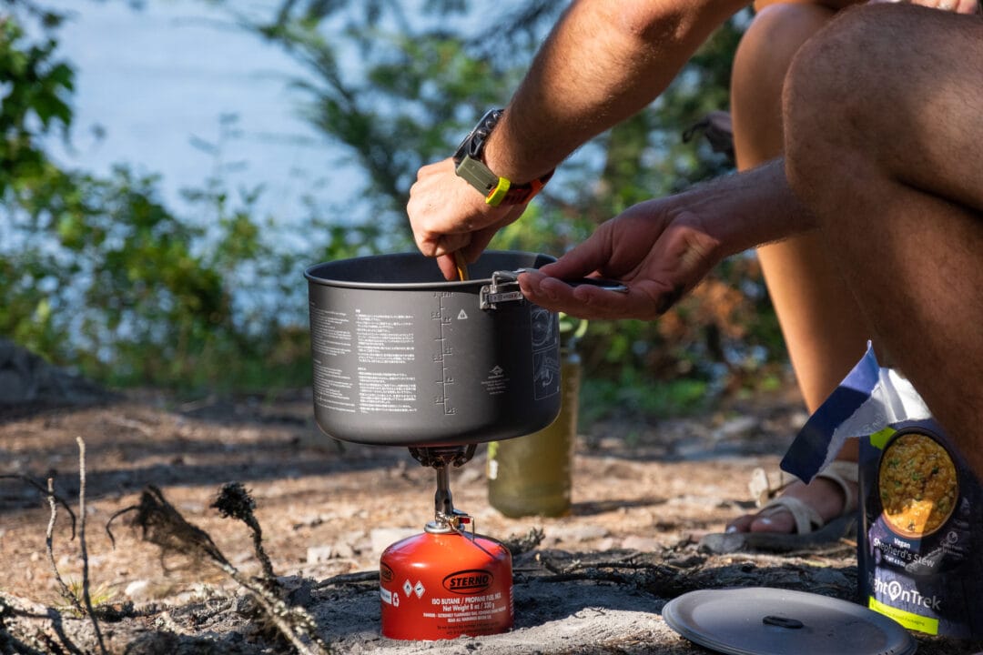 A person's hands making a campground meal in a camp kitchen with a view of water in the background