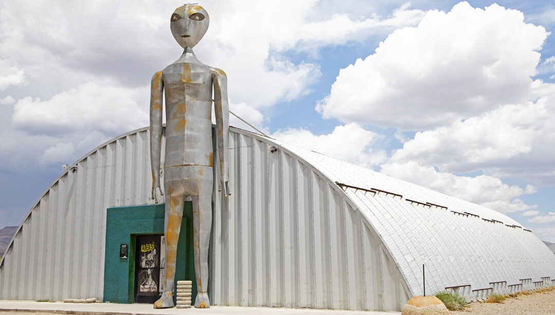 A tall silver alien statue stand outside of a building in the desert