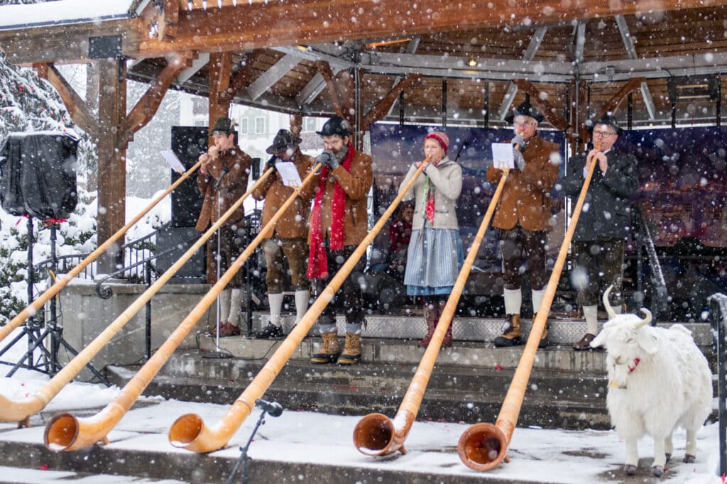 A group of musicians wearing traditional German attire and playing a long horn instrument in the snowfall