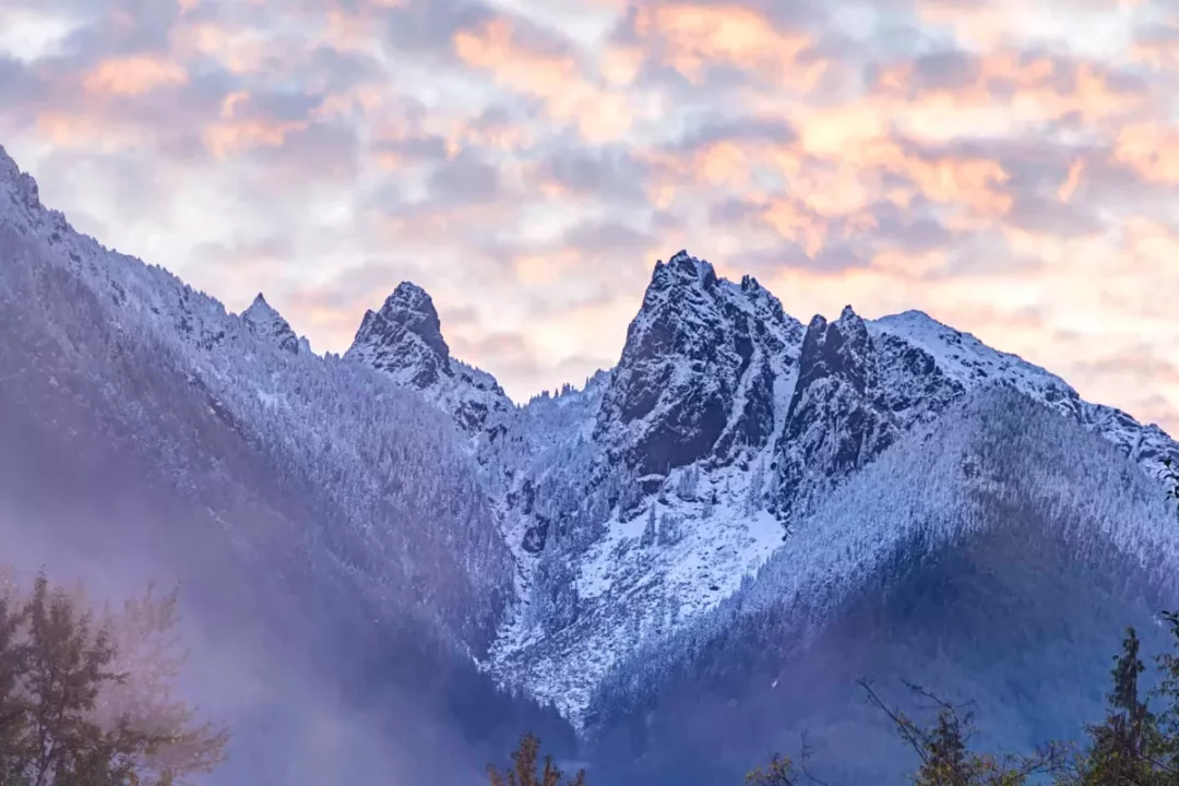 Craggy mountain peaks are covered with snow with a colorful sky full of clouds as the backdrop.