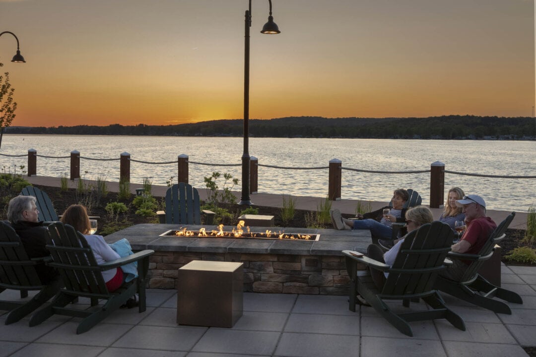A waterside firepit burns bright as the sun sets in the distance.