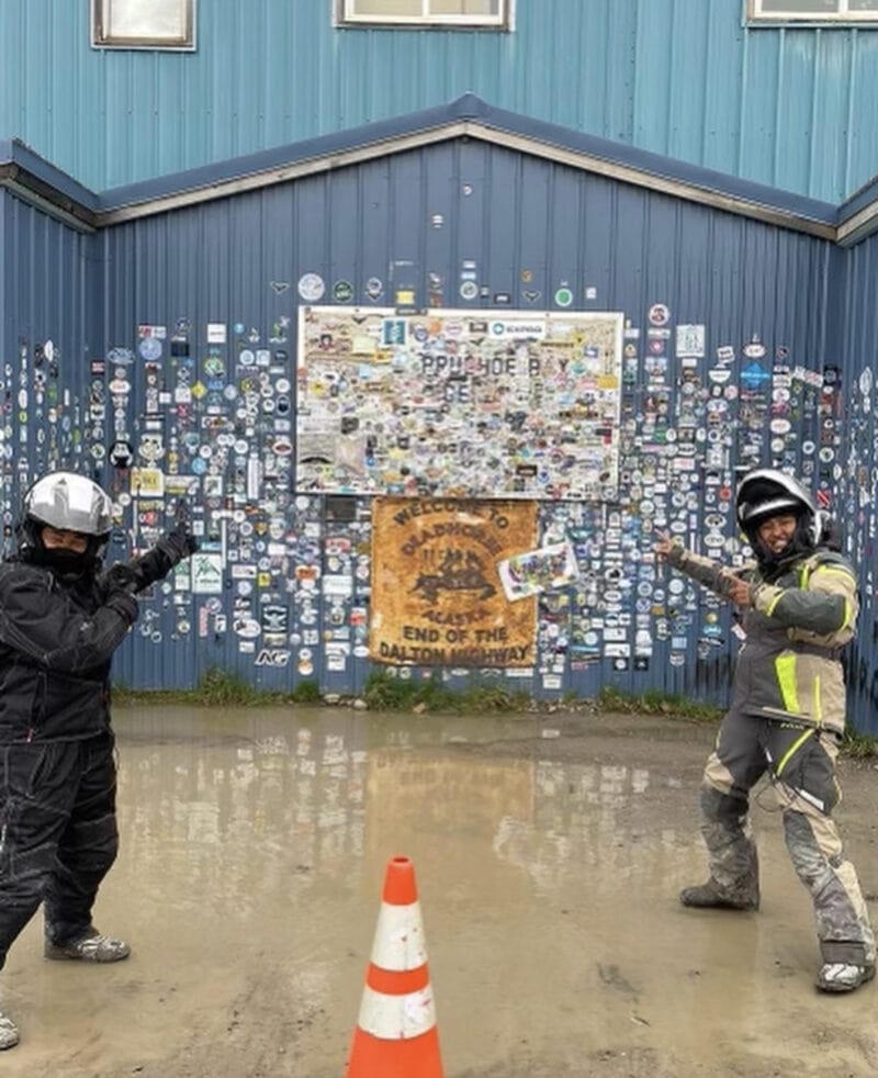 Two women in motorcycle gear pointing to a sign for Prudhoe Bay, covered in stickers