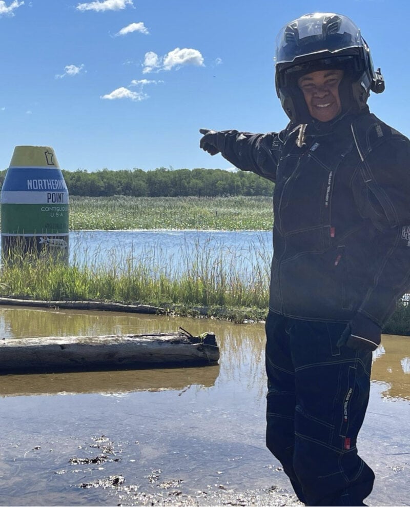 A woman in motorcycle gear stands next to a small lake and points to a marker for the Northernmost Point in the Contiguous U.S.