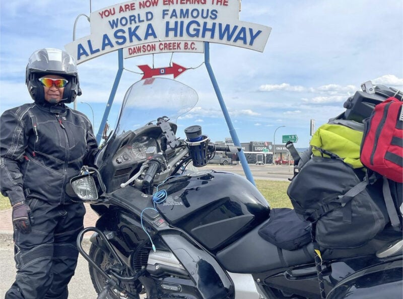 A woman in motorcycle gear standing next to a motorcycle at a sign for the World-Famous Alaska Highway