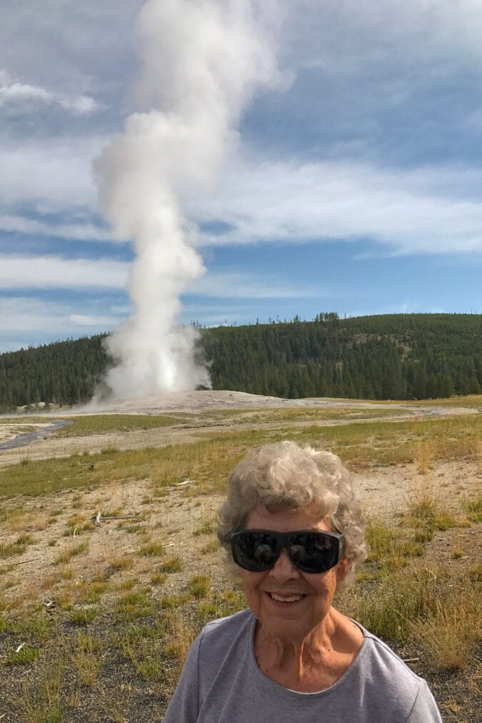 A geyser erupts in the background as a woman smiles for a photo in the foreground.