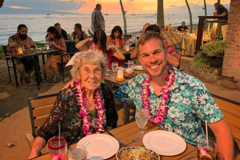 A grandmother and her adult grandson wear leis as an ocean sunset appears in the background.