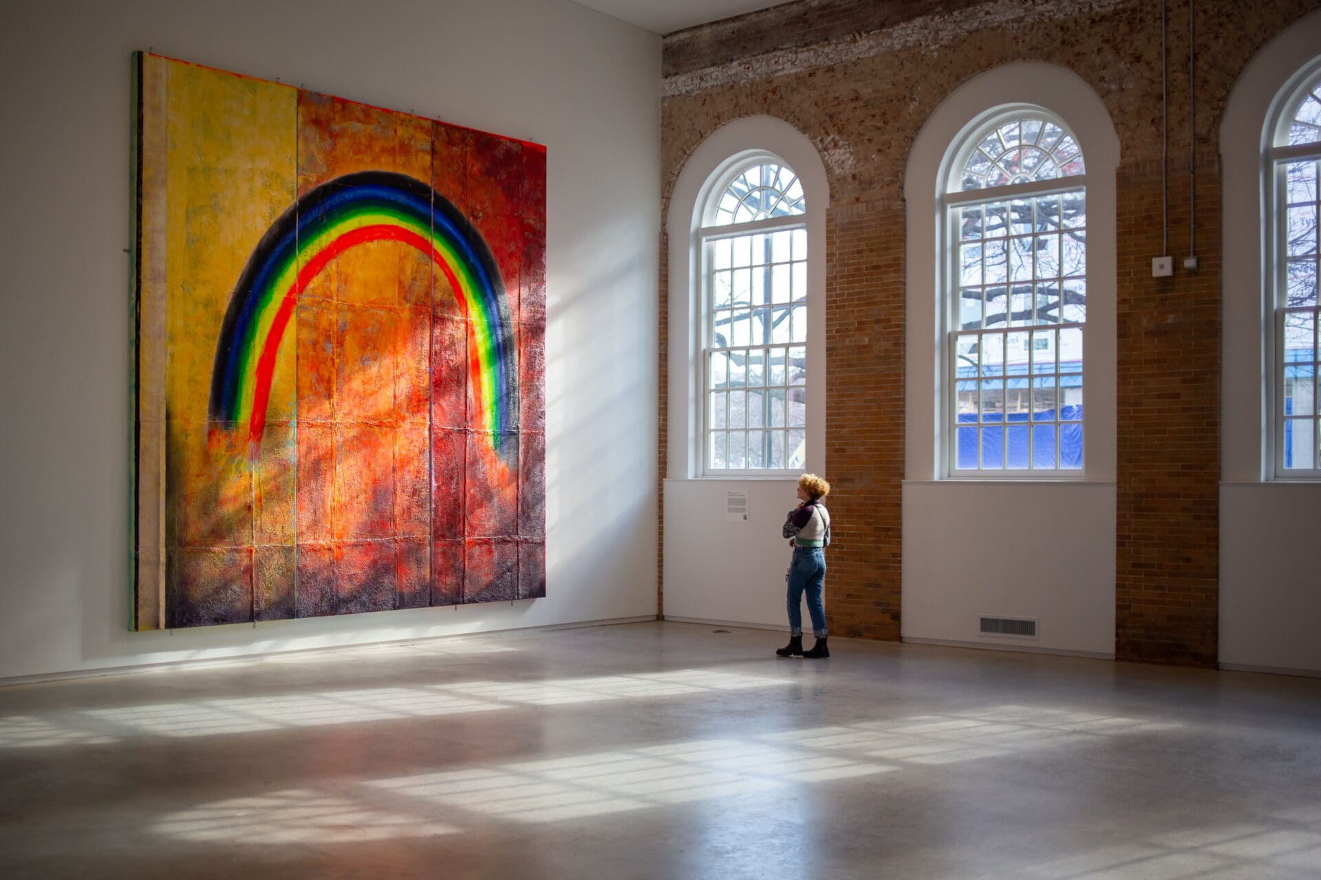 a person stands in a large art gallery looking at a painting of a rainbow while sunlight streams in through the arched windows
