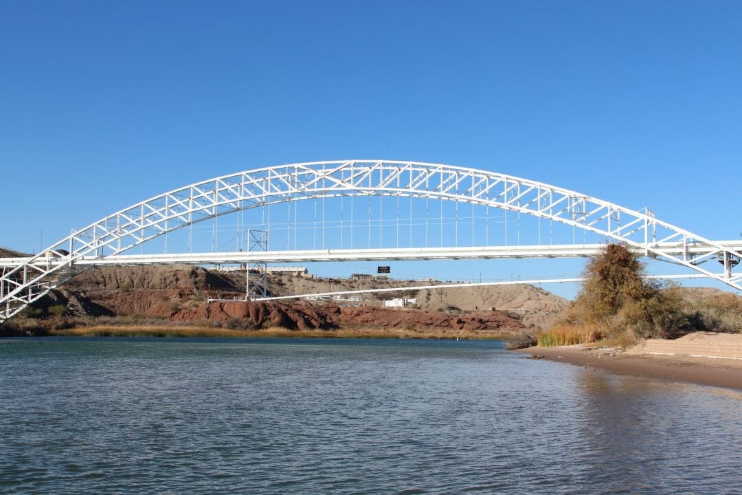 a large white arched steel bridge stretches over water against a clear blue sky