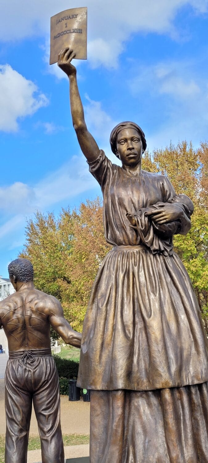 A bronze statue of a Black woman holding a baby in one arm and a document in the other. A man with scars across his body has his back turned against her. 