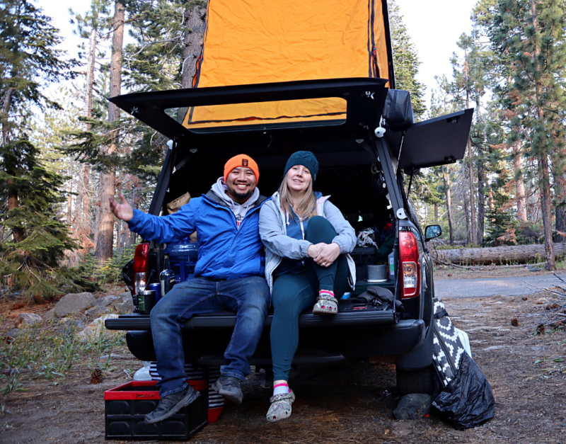 A man and woman sit in a truck bed with a camper attached to the top.