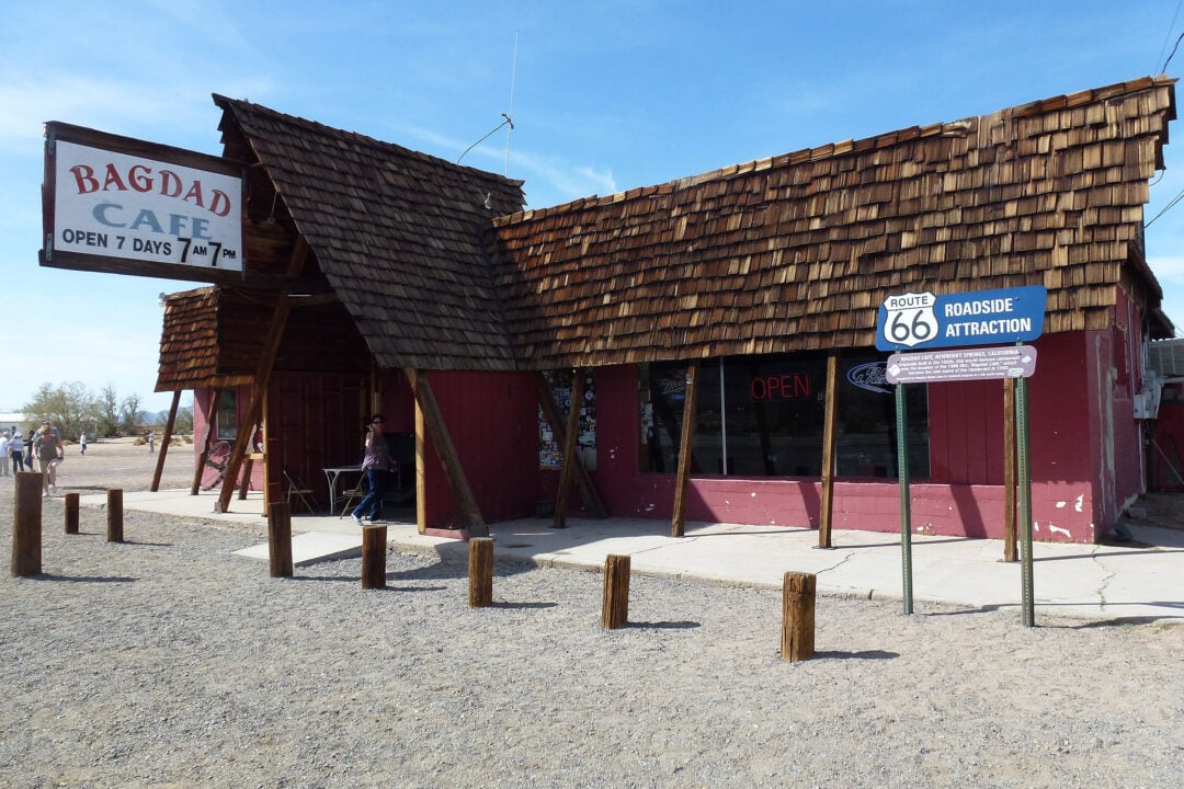 a small red building with a wooden shingle roof in the desert with a sign that says "bagdad cafe open 7 days 7am 7pm"