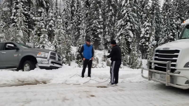 Two men stand on a snowy roadway holding jumper cables.