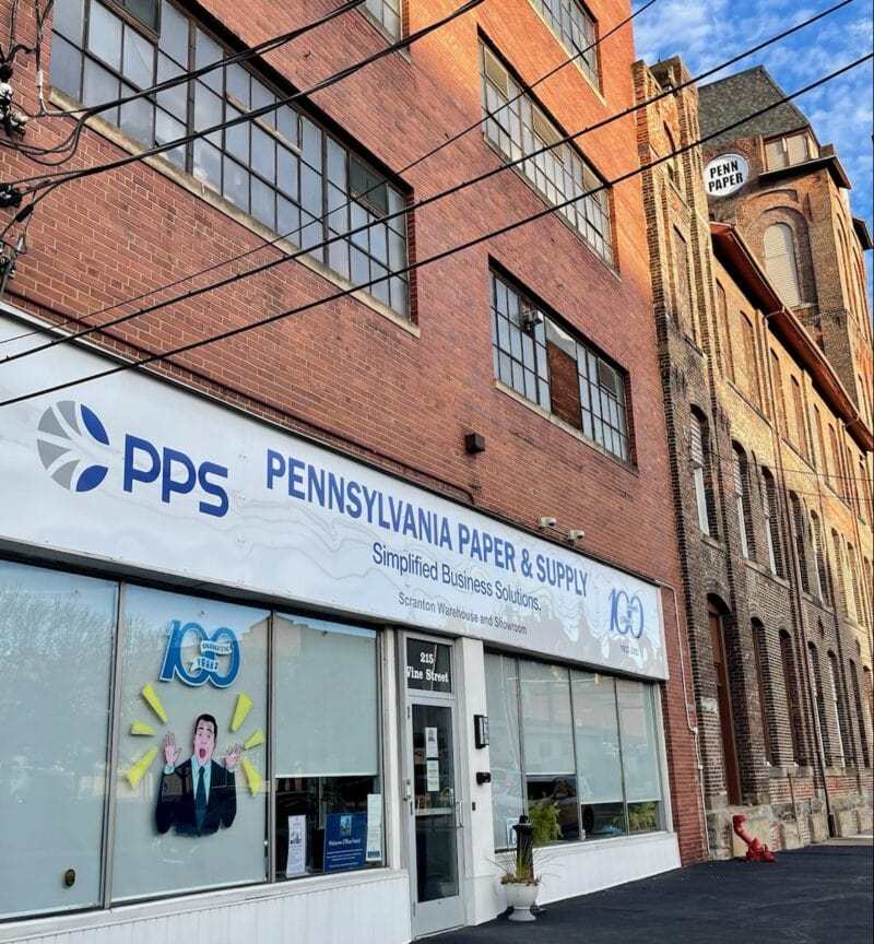 The storefront of a paper company stands in the foreground with a large brick tower standing in the background.