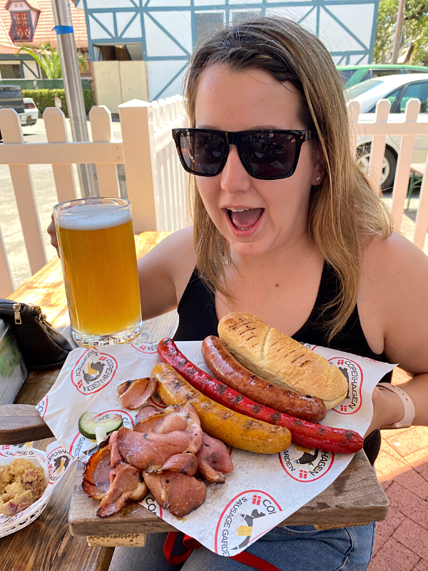 A woman looks on at delight at a meal that includes a massive sausage and beer.