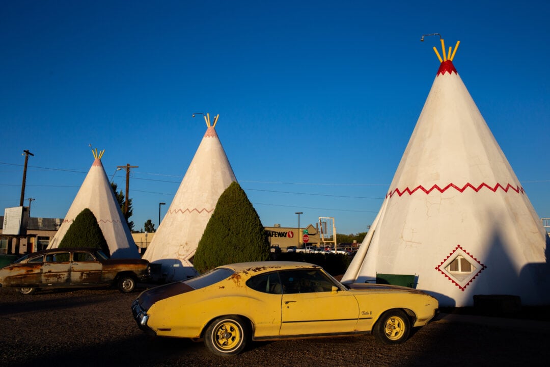three concrete teepees set against a blue sky with a vintage yellow car parked out front