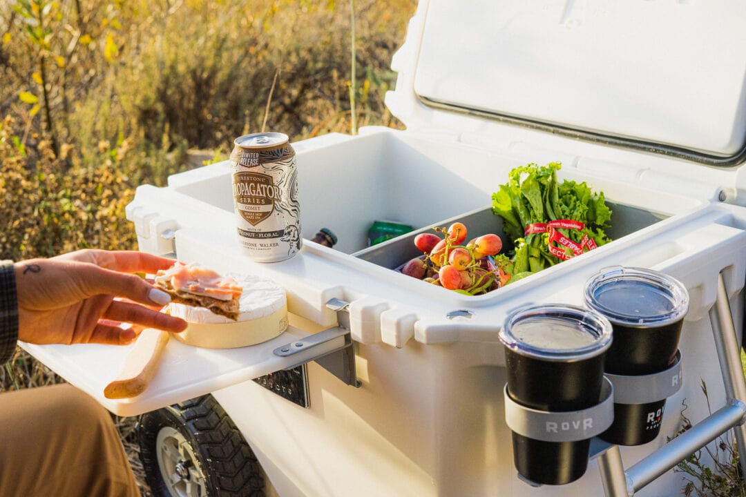 Hard-sided cooler is open in a field with drinks and food inside. Outside there is a cutting board attachment with wheel of cheese and two cup holders.