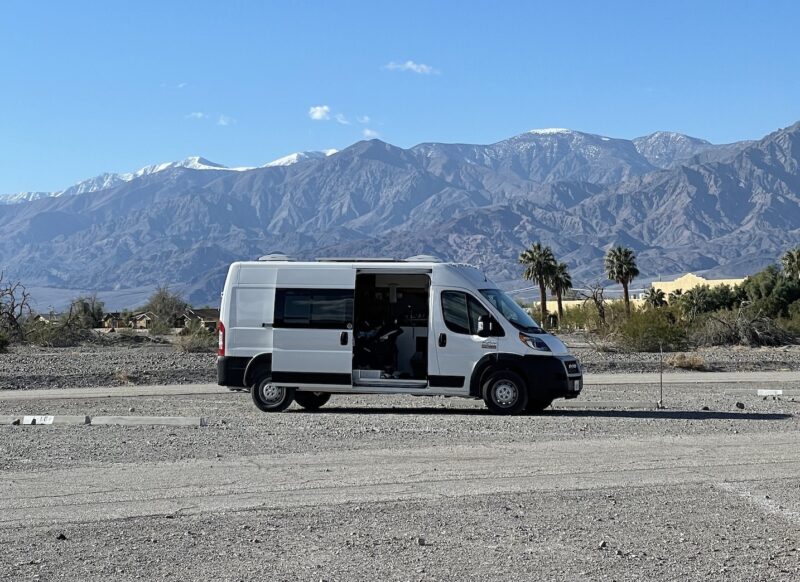 A white campervan is parked in a gravel parking lot with palm trees and snow-capped mountains in the background