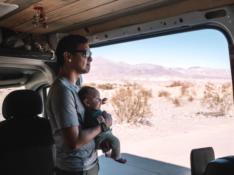 Dad holding a baby inside a campervan looking out onto desert landscape from an open door