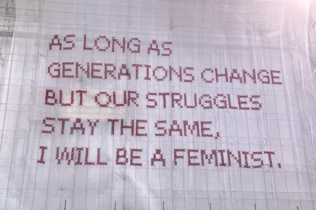 white netting draped over a building stitched with pink netting that says “As long as generations change but our struggles stay the same, I will be a feminist.”