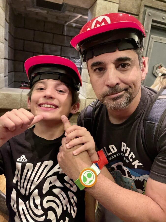 A father and son don matching Mario hats as they wait to compete on an AR Mario Kart track