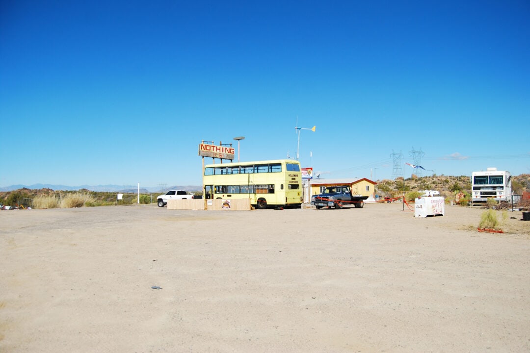 a desert encampment including a few vehicles, a double decker yellow bus and a sign that says "nothing"