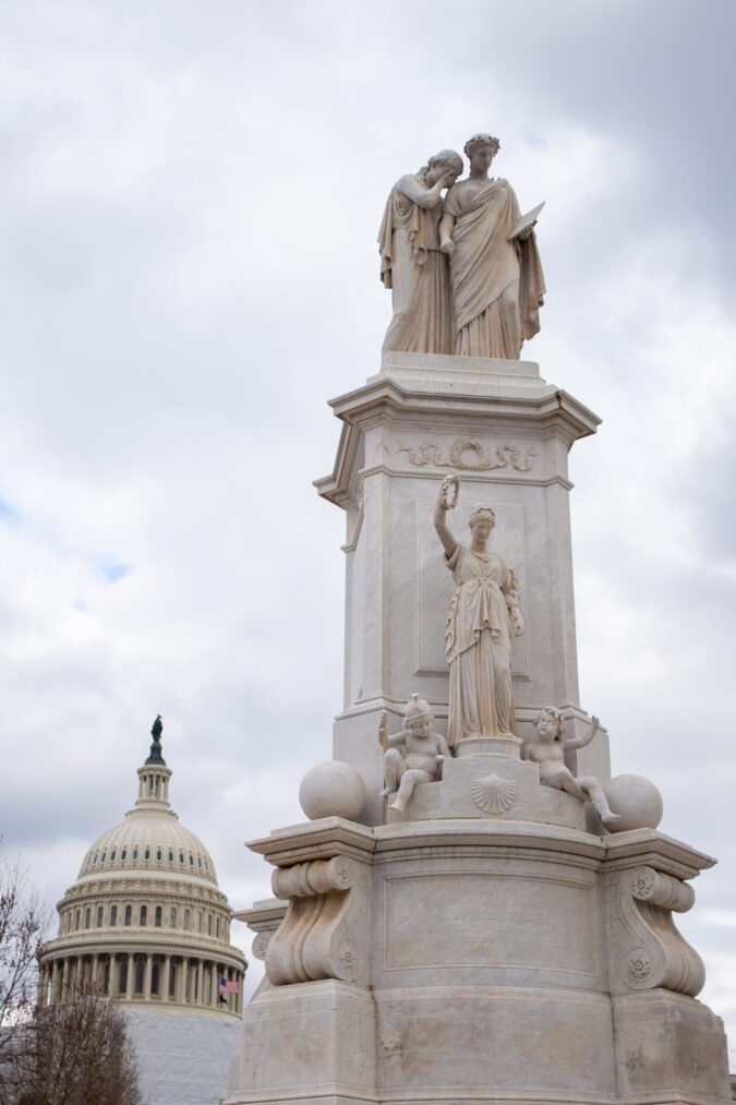 a white marble monument featuring classical female figures stands in front of the capitol dome