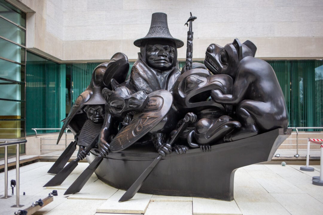 a black bronze sculpture of mythical figures in a canoe outside in the plaza of the canadian embassy