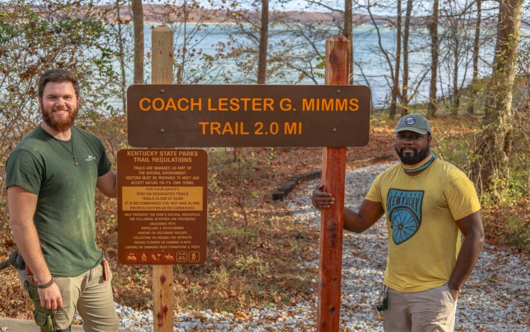 Two men stand by a sign that reads "Coach Lester G. Mimms Trail 2.0 Miles"