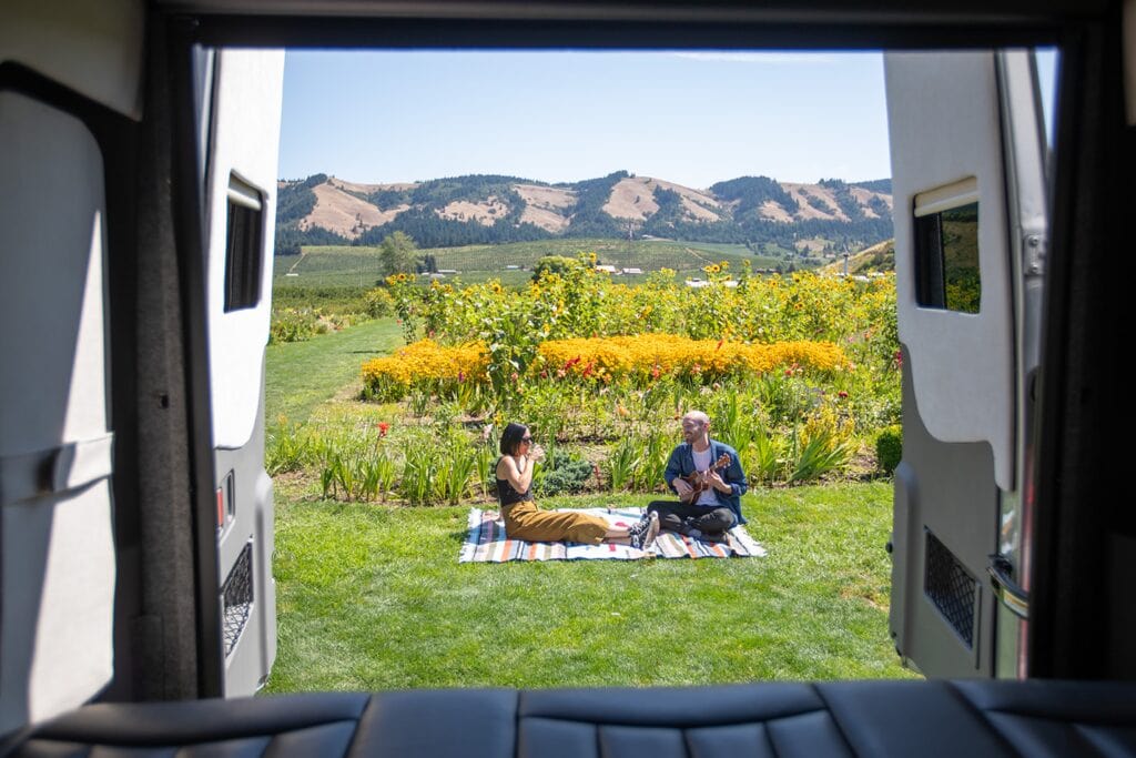 The back doors of a Class B camper are open to reveal a couple enjoying a picnic.