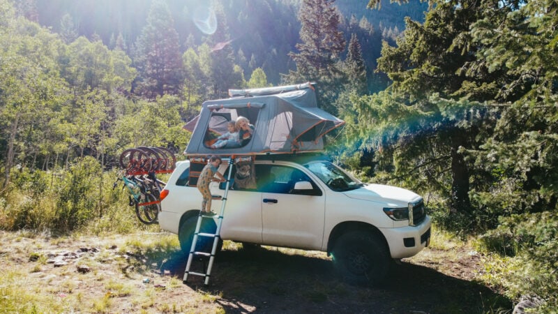 Large white SUV at a campsite with a rooftop tent and kids looking out from inside