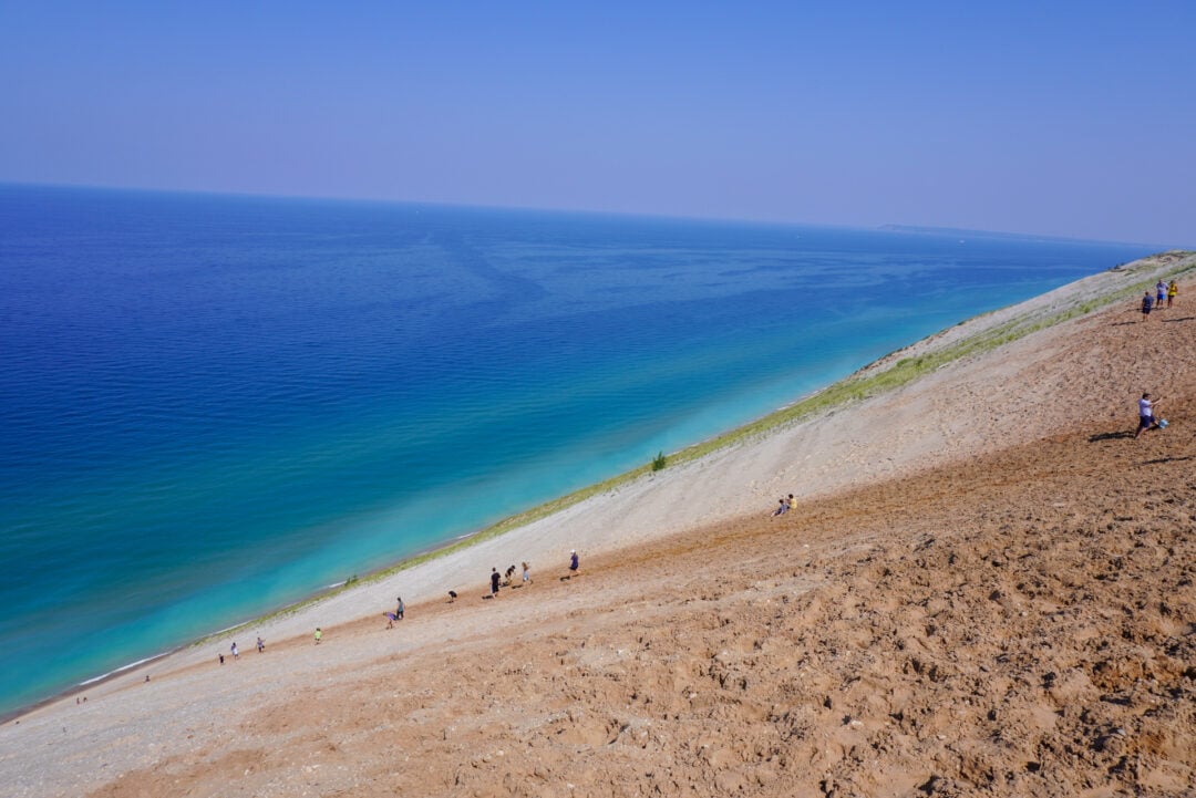 A steep sand dune leading down into clear blue water. People are hiking up and down the large dune.