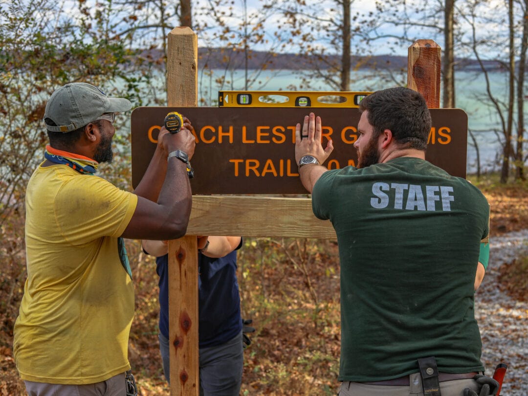 Two men use power tools to hang a sign for a hiking trailhead
