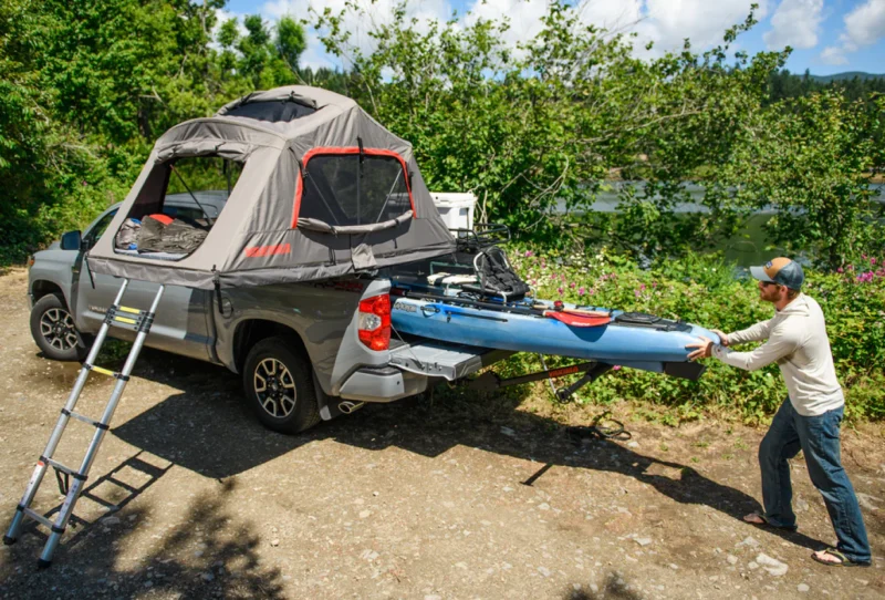 Man loading a kayak in the back of a truck with a rooftop tent on the bed