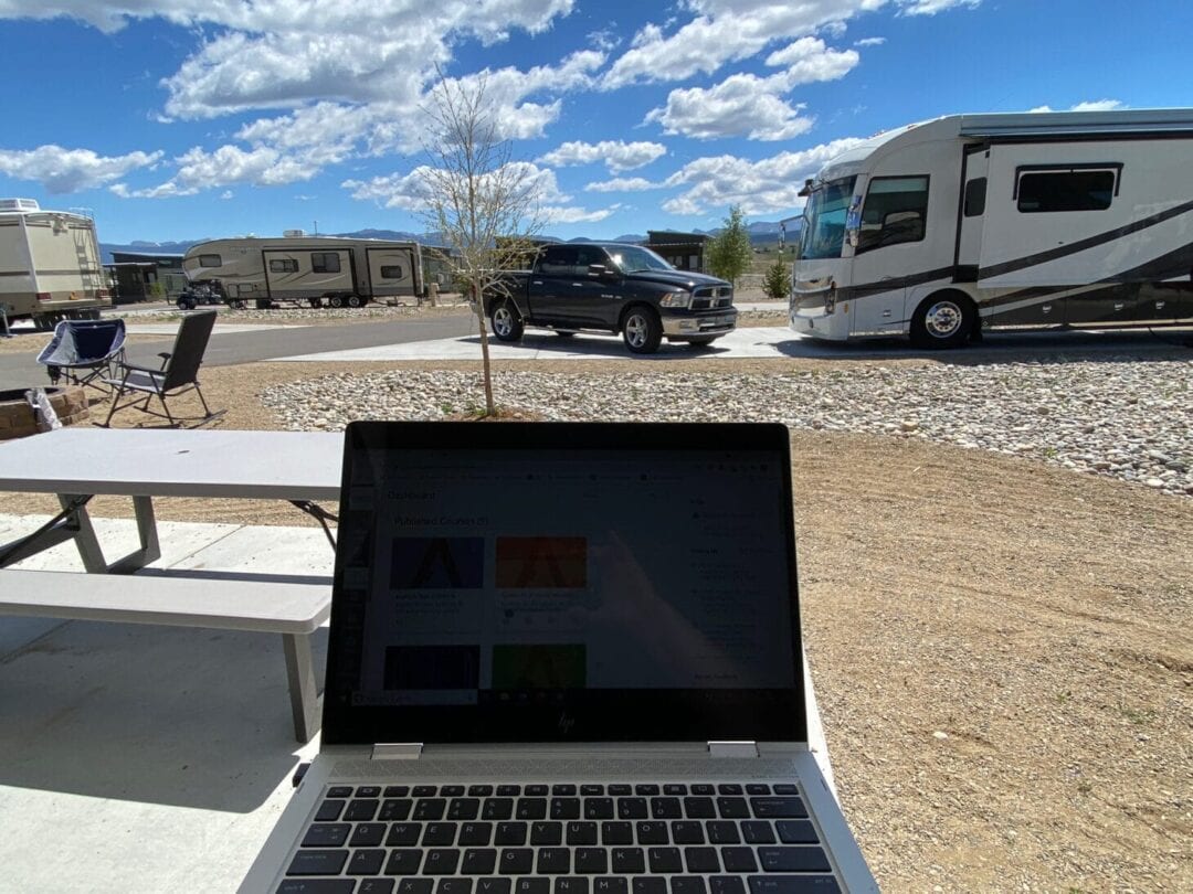 Laptop open with work screen sitting on table outside at a campground with RVs surrounding the campsite