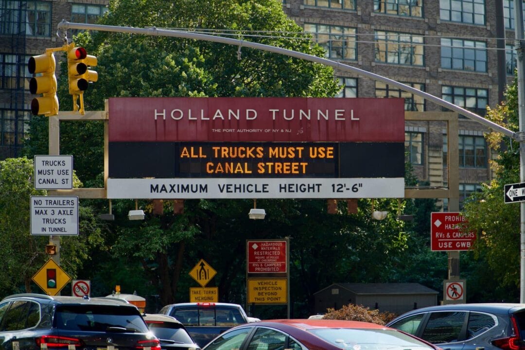Sign on busy street that reads "Holland Tunnel: All trucks must use Canal Street. Maximum vehicle height 12 feet, 6 inches"