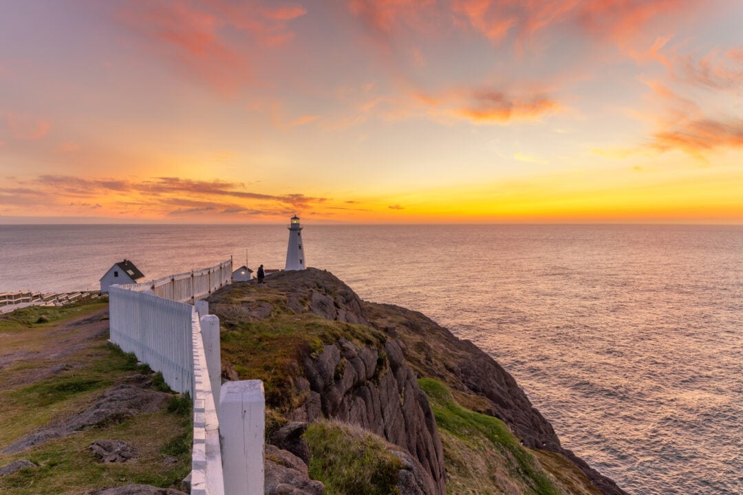 A small lighthouse sits at the end of a rocky cliff, surrounded by pink skies