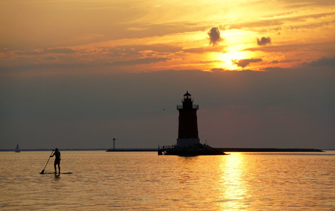 a silhouette of a person paddle boarding in front of a lighthouse at sunset
