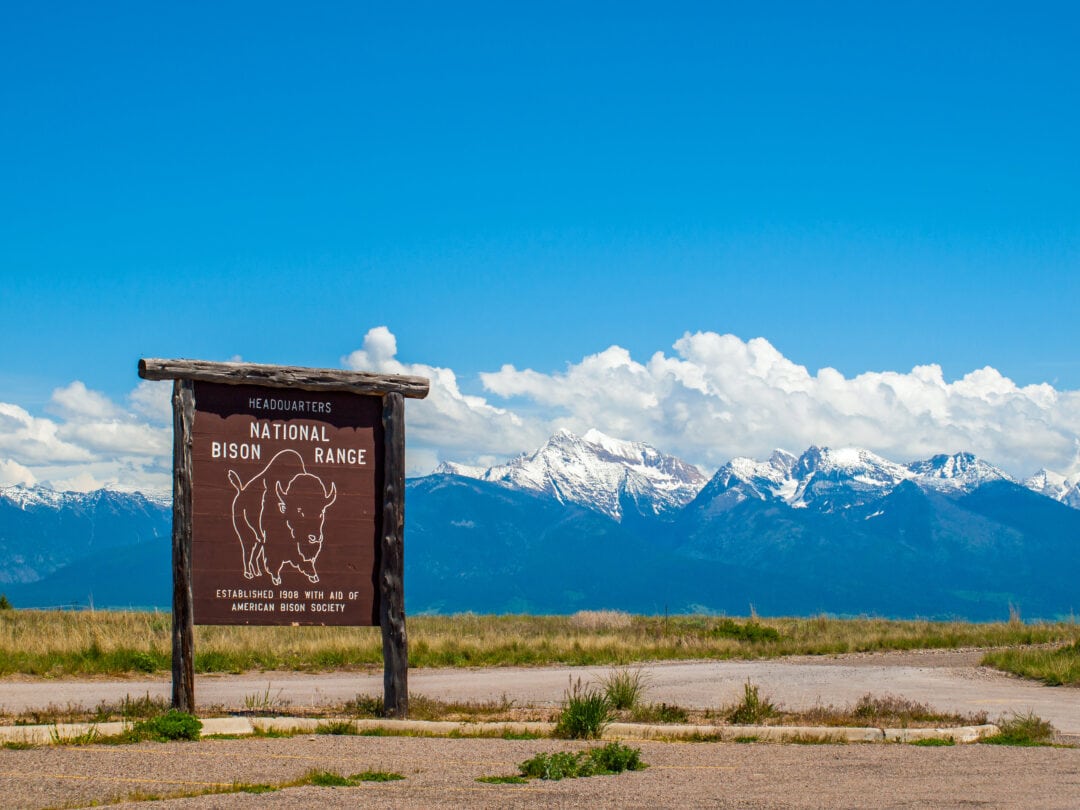 a wooden sign for the national bison range stands in front of a snowy mountain range