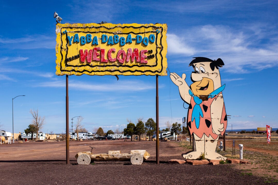 a large wooden sign in front of a campground featuring a cutout of fred flintstone pointing to a yellow sign with the letters "yabba-dabba-doo welcome"